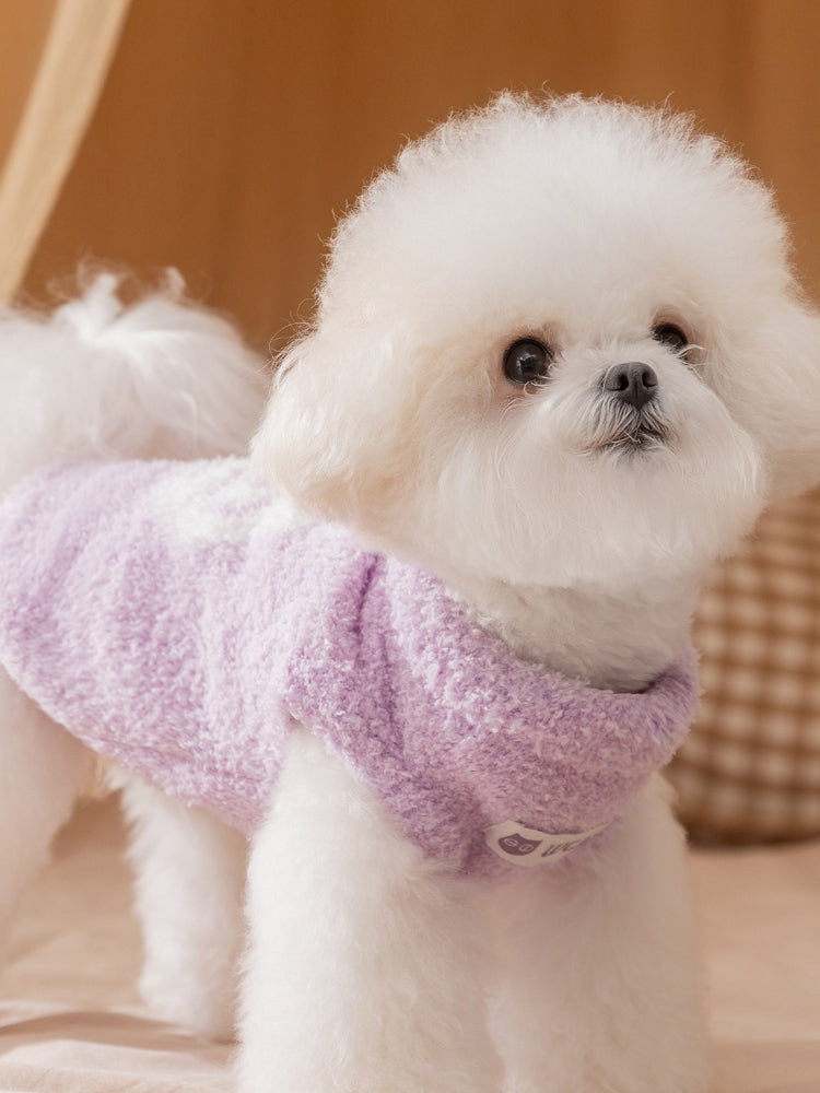 White bichon frise dog wearing a lavender fluffy dog sweater with a white "Wcat" logo patch on the chest.