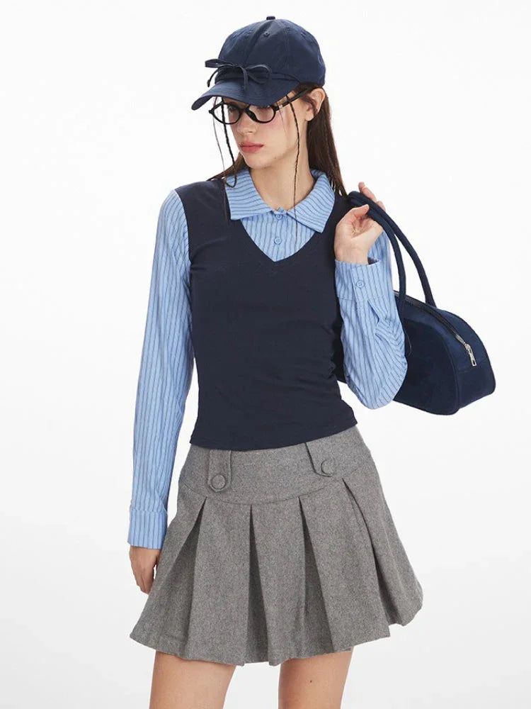 Close-up of a woman wearing a blue striped collared shirt, navy V-neck vest, and grey pleated skirt, with glasses and a navy baseball cap.