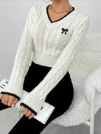 Woman sitting while wearing a white cable knit V-neck cropped sweater with black trim and holding a book.