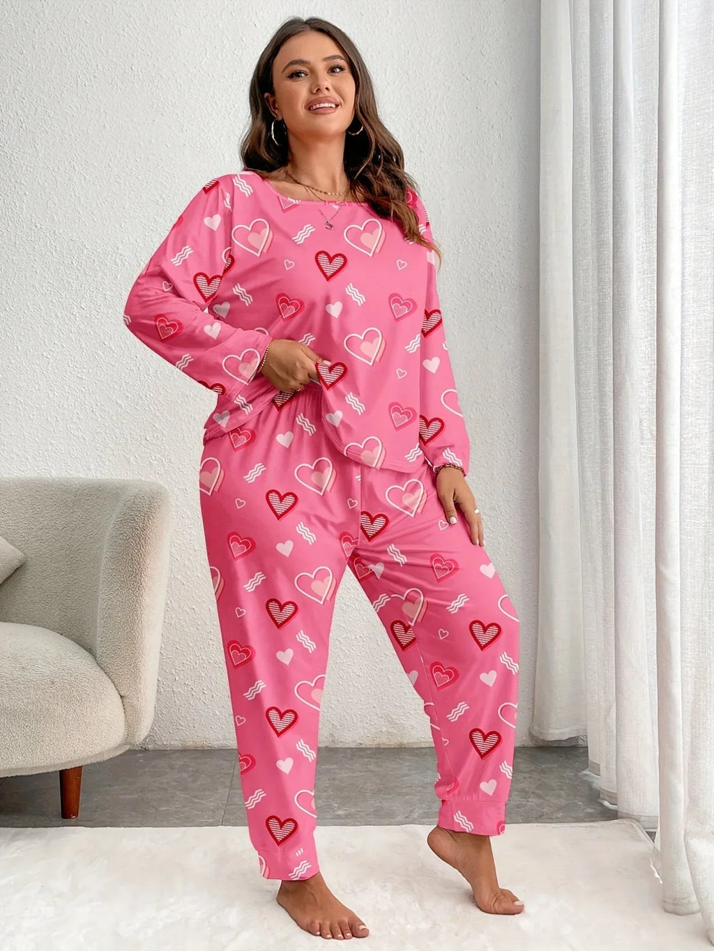 Woman wearing a pink long-sleeved pajama set with an all-over heart print, standing and adjusting her top.