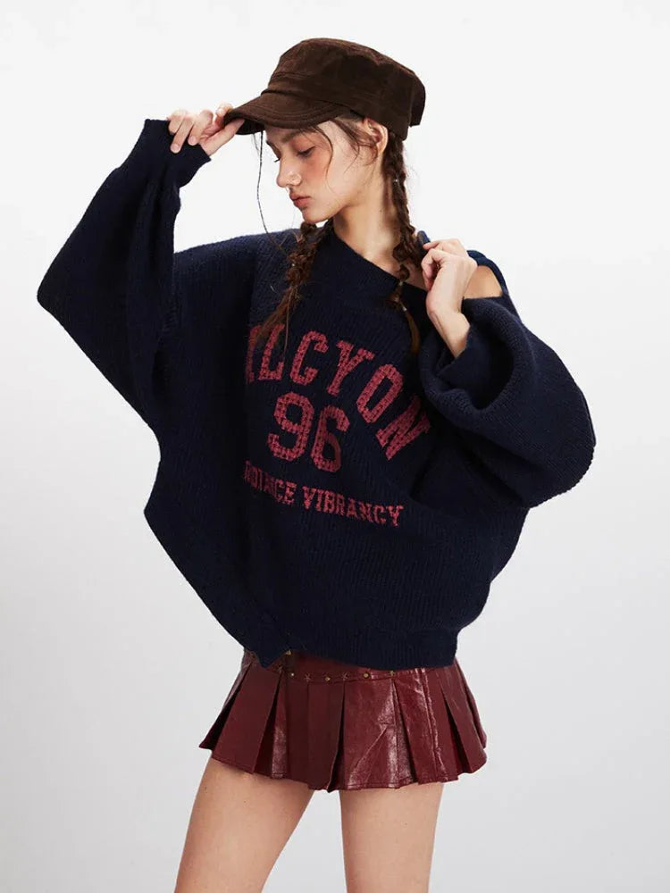 Woman in a navy blue oversized knit sweater with dropped shoulders and wide sleeves, styled with a burgundy faux leather mini skirt and brown cap.