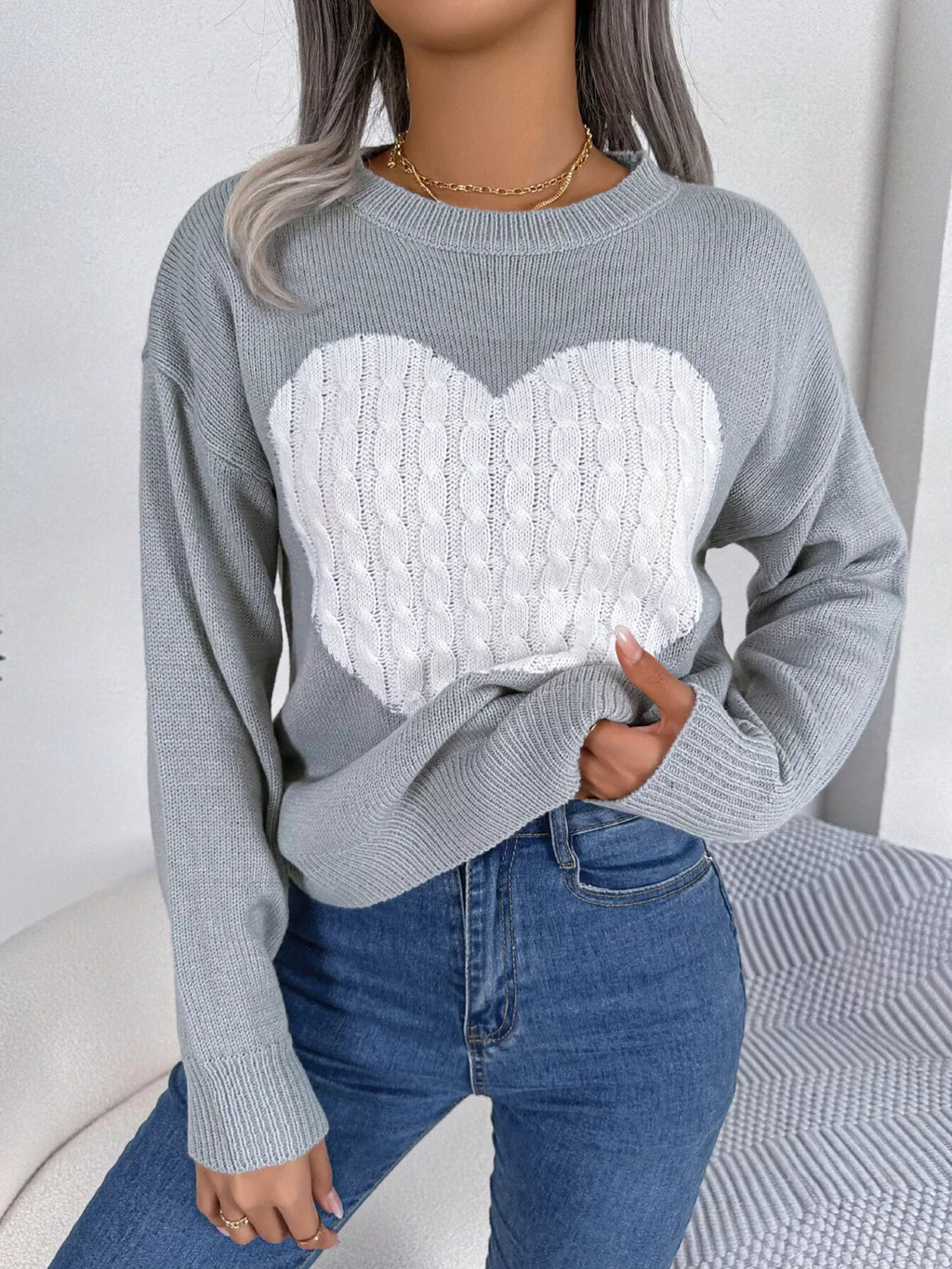 Woman wearing a grey long sleeve knit sweater with a large white cable knit heart graphic on the front.