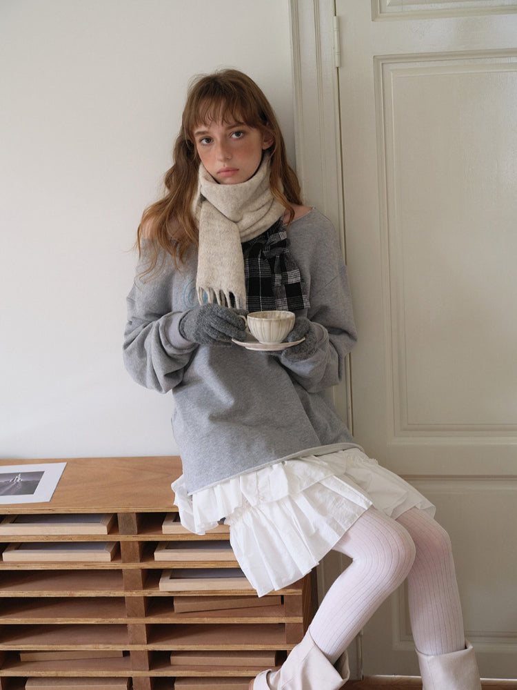 Woman wearing a grey sweatshirt, white ruffled skirt, light pink ribbed tights, and a cream scarf with fringe detail