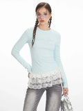 Woman posing in a light blue long-sleeved top with white lace trim and gray jeans.