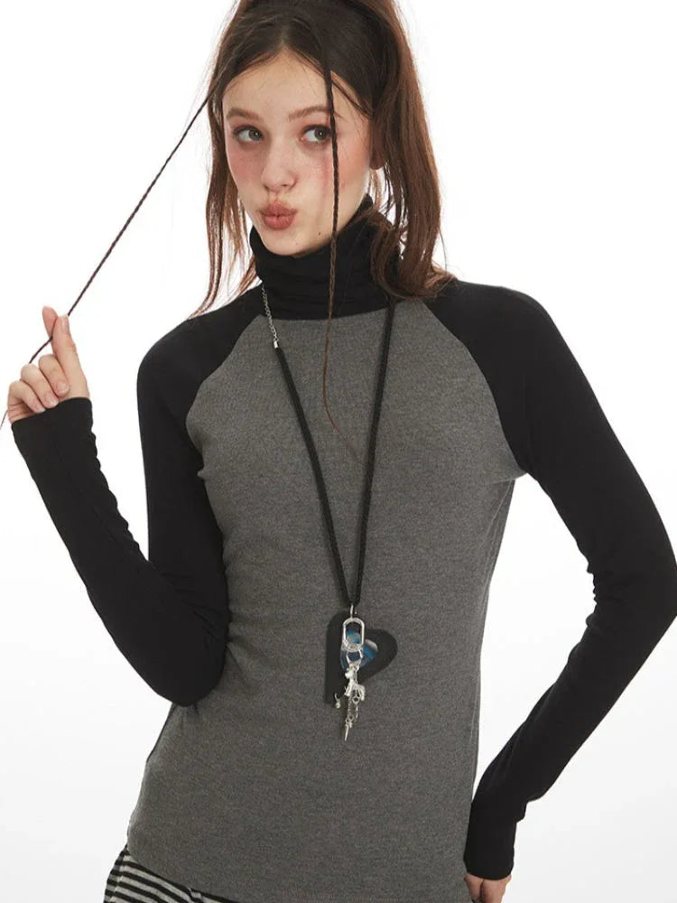Close-up of a woman's face and upper torso wearing a grey long-sleeved turtleneck top with black raglan sleeves and a pendant necklace.
