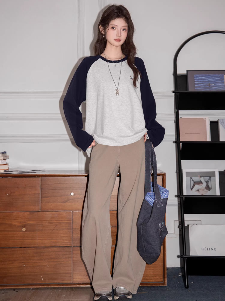 Full-body shot of a woman wearing a light grey long-sleeve raglan shirt with navy blue sleeves, khaki wide-leg pants, and holding a tote bag.