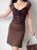 Full body shot of a woman in a brown floral mesh top and a matching brown skirt, holding a brown handbag.