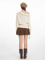 Back view of a model wearing a cream-colored off-the-shoulder knit sweater with a diamond pattern and decorative trim. The sweater is paired with a brown textured mini skirt and brown knee-high socks.