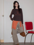 Woman leaning against a wall wearing a dark brown long sleeve top with embroidery.