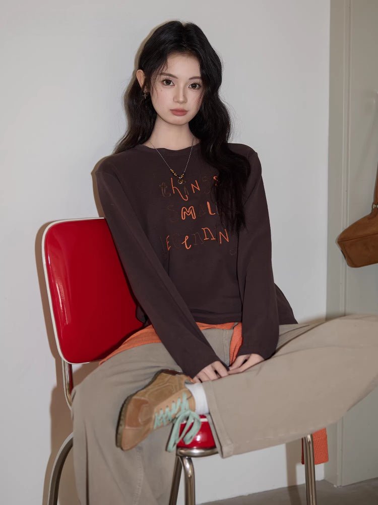 Woman sitting on a red chair wearing a dark brown long sleeve top with orange embroidery.