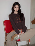 Woman sitting on a red chair wearing a dark brown long sleeve top with orange embroidery.