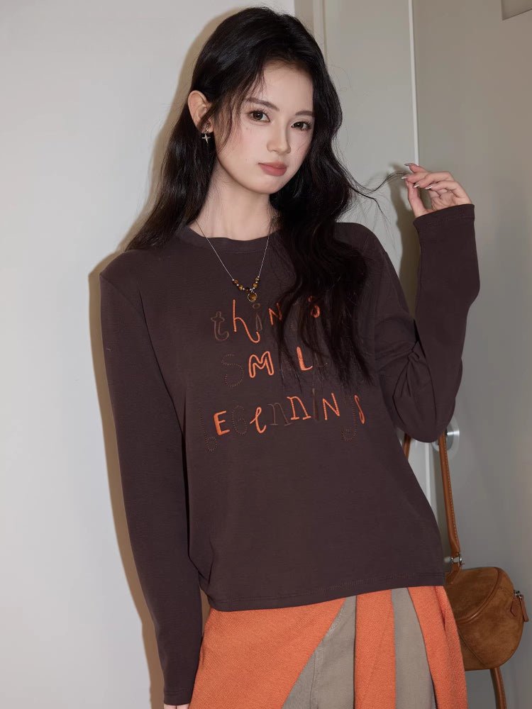 Close-up of a dark brown long sleeve top with contrasting orange letter embroidery.