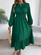 Full view of a woman in a green A-line dress with puff sleeves and a ruffled hem, holding a straw bag.