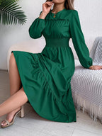 Woman sitting on a sofa wearing a long-sleeved green A-line dress.