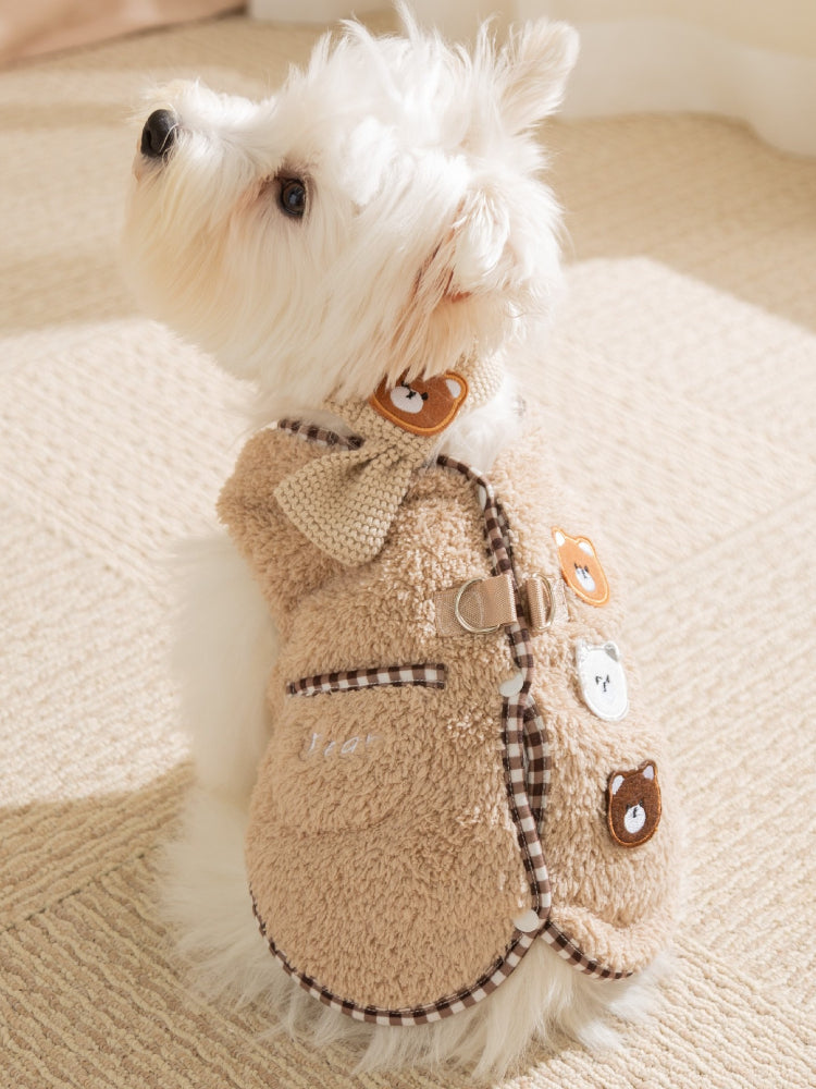 Small white dog wearing a tan fleece vest with bear appliques and plaid trim.