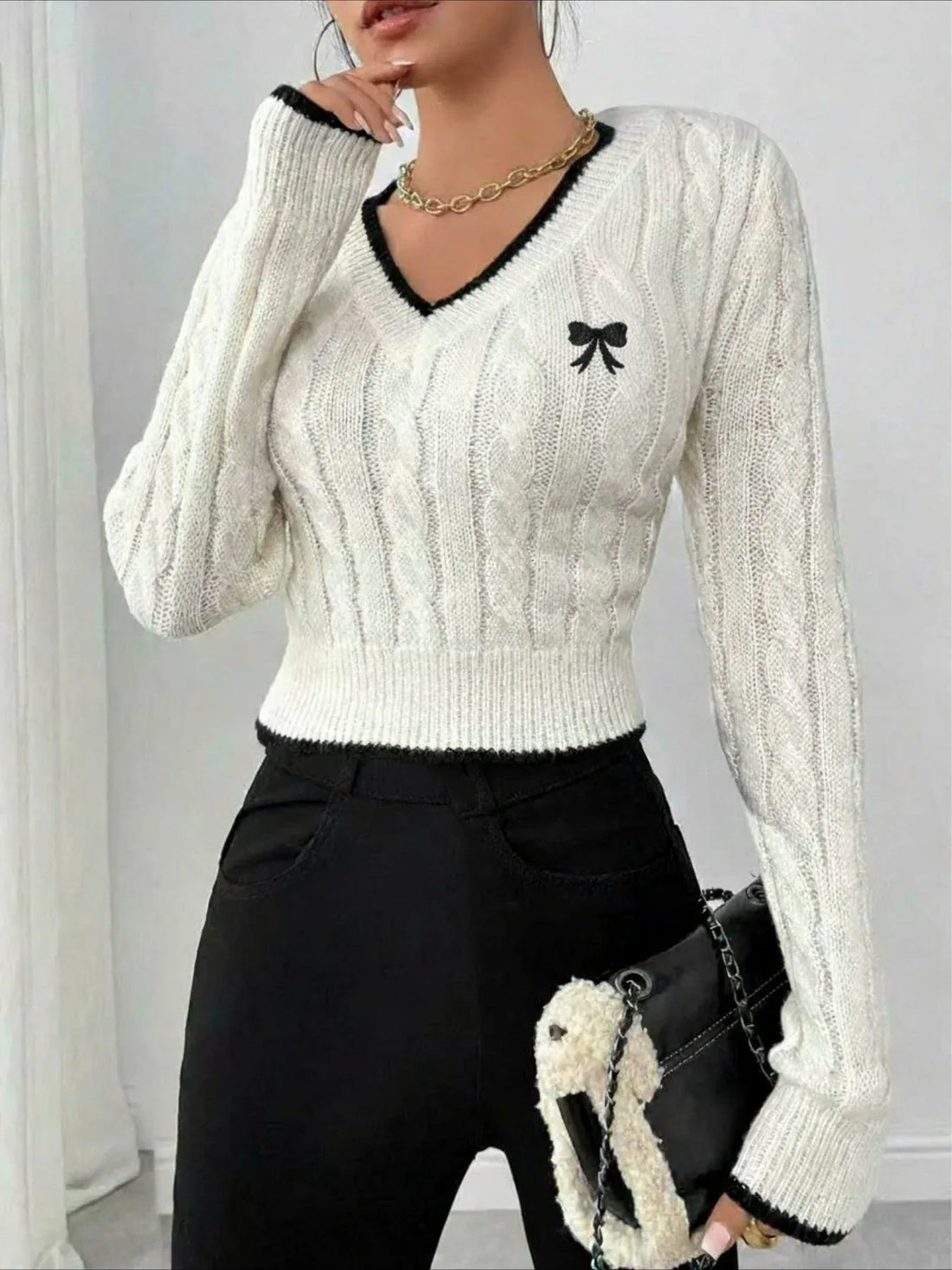 Woman wearing a white cable knit V-neck cropped sweater with black trim and an embroidered bow detail.