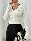 Woman wearing a white cable knit V-neck cropped sweater with black trim and an embroidered bow detail.