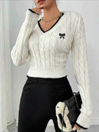 Woman wearing a white cable knit V-neck cropped sweater with black trim and an embroidered bow detail.