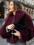 Woman wearing a burgundy shaggy faux fur cropped jacket and grey sweater, holding a black clutch bag.