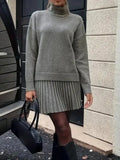 Woman in a grey knit turtleneck sweater and pleated mini skirt standing next to a car door, holding a black handbag.