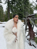 Woman wearing a long white faux fur coat over a white bikini in a snowy forest