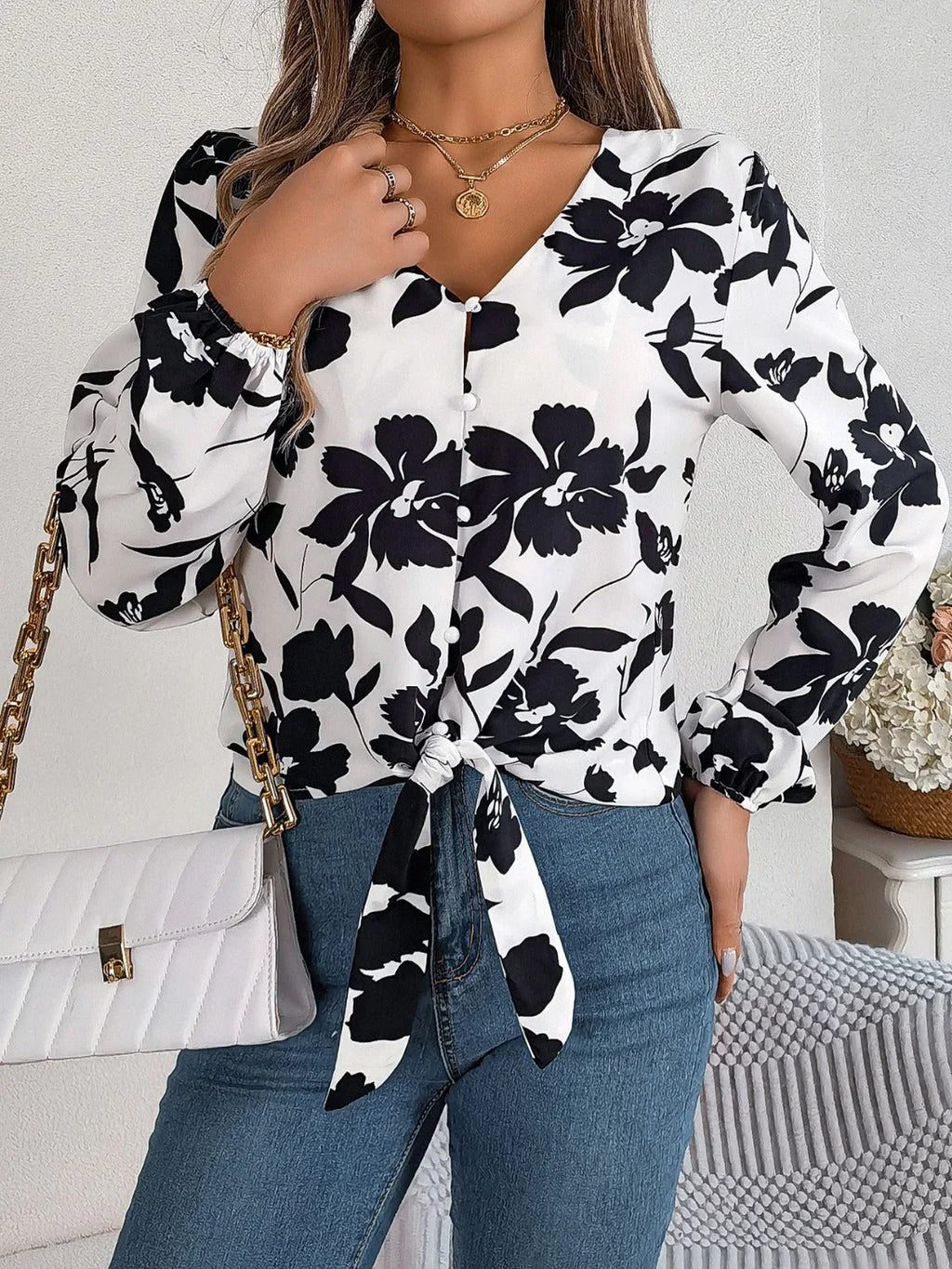 Woman wearing a white long-sleeve blouse with a black floral print and a tie-front detail, holding a white quilted handbag with a gold chain strap.