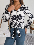 Woman wearing a white long-sleeve blouse with a black floral print and a tie-front detail, holding a white quilted handbag with a gold chain strap.