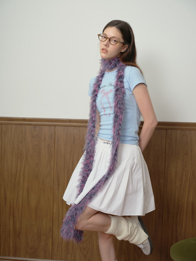 Woman wearing a light blue graphic t-shirt with a pink bow print, a white pleated mini skirt, a long purple and pink faux fur scarf, cream leg warmers, and grey sneakers.