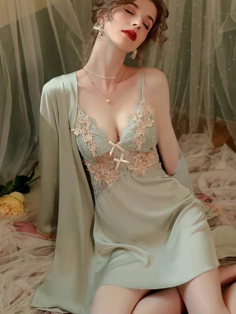 Woman wearing a light green satin slip dress with lace detailing and a matching robe, seated on a bed.