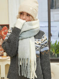 Woman wearing a knit sweater with a geometric pattern, a multi-colored scarf, and a beige beanie, covering her mouth with her gloved hand.