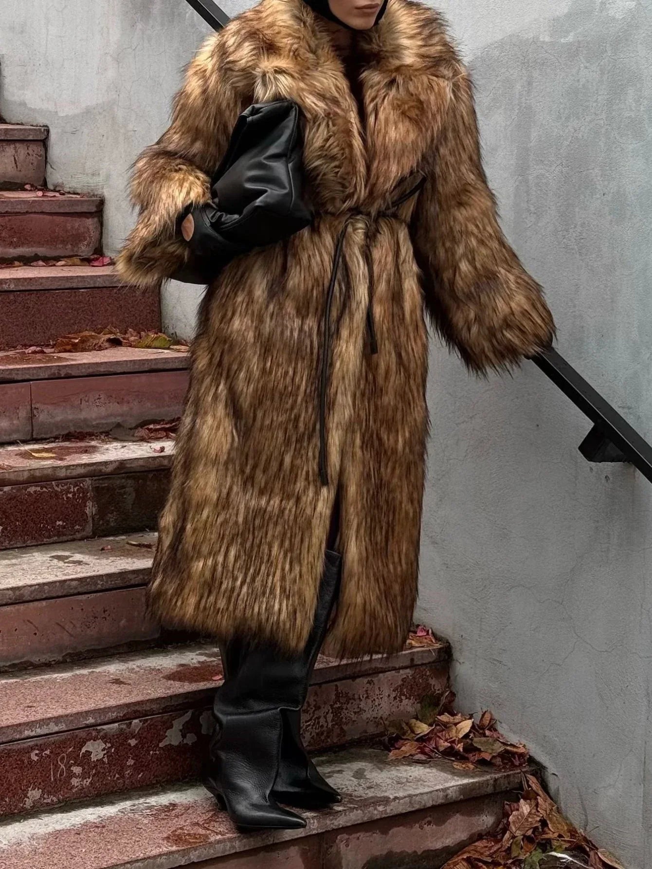 Woman wearing a long brown faux fur coat with a belt, black boots, and a black handbag.