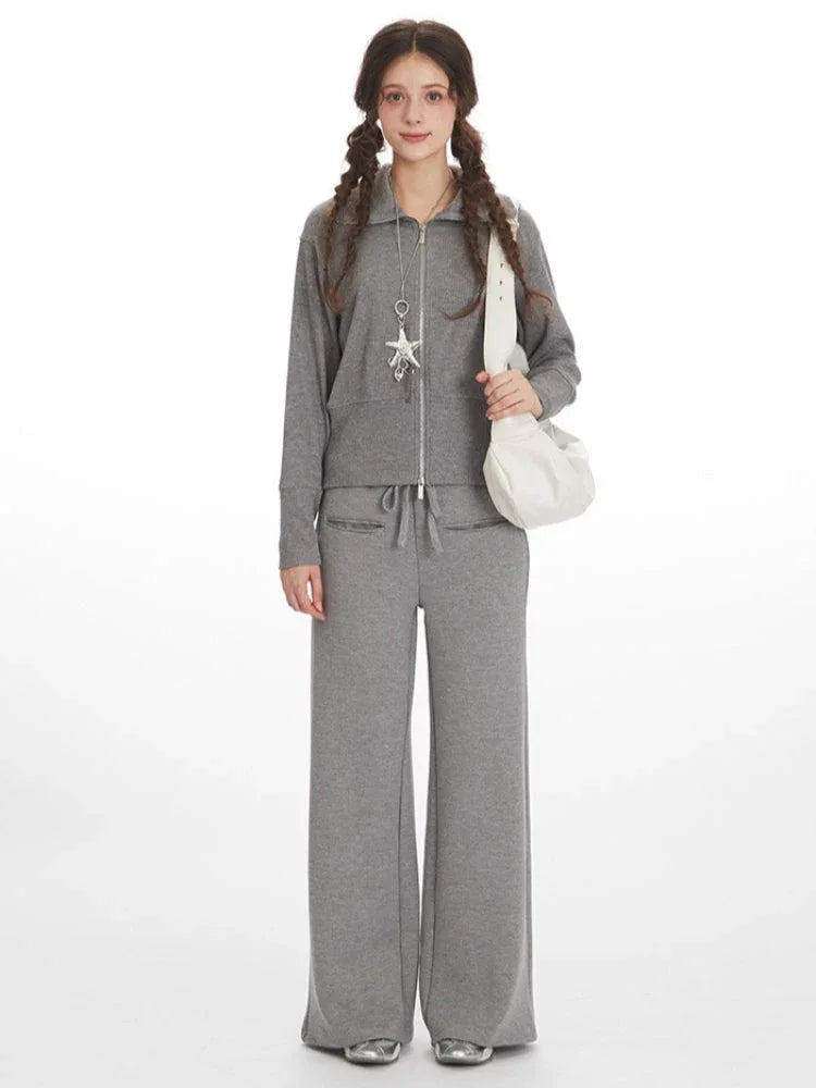 Woman wearing a grey ribbed knit loungewear set with a full zip jacket and wide leg pants, carrying a white shoulder bag. Main image