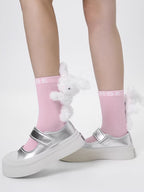 Pink crew socks with a plush white bunny charm and 'MSE' logo, worn with silver Mary Jane platform shoes.