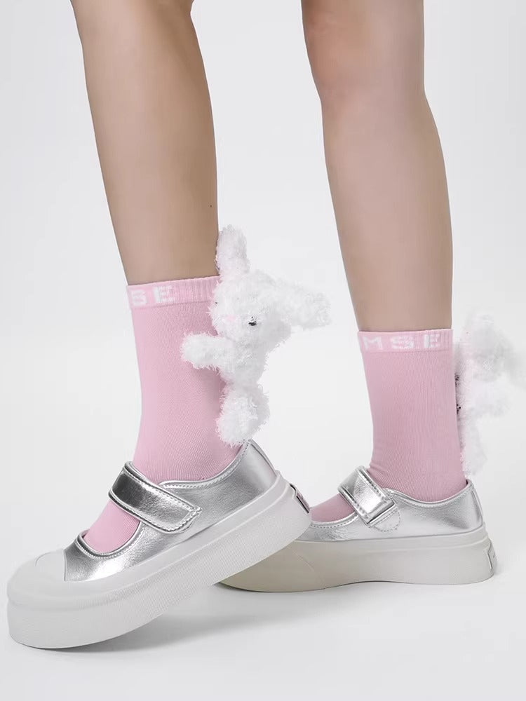 Pink crew socks with a plush white bunny charm and 'MSE' logo, worn with silver Mary Jane platform shoes.