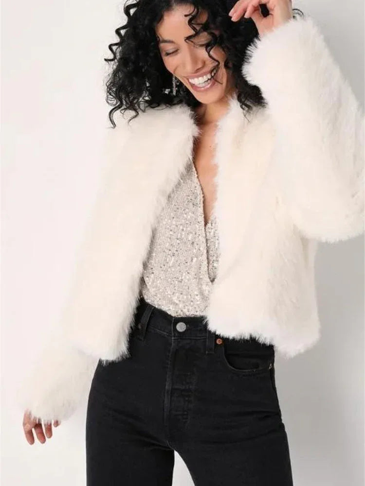 Woman smiling, wearing a white faux fur cropped jacket over a sparkly top and black jeans.