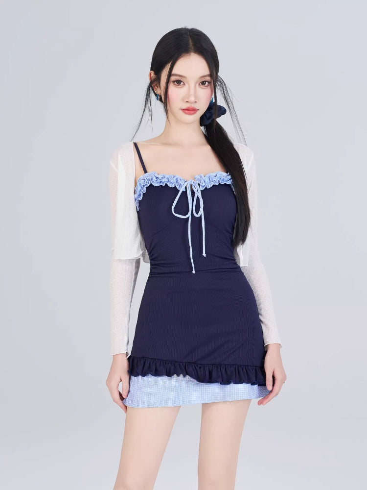 Woman in navy one-piece swimsuit with blue gingham skirt overlay, white sheer cardigan, and hair tied with blue ribbon