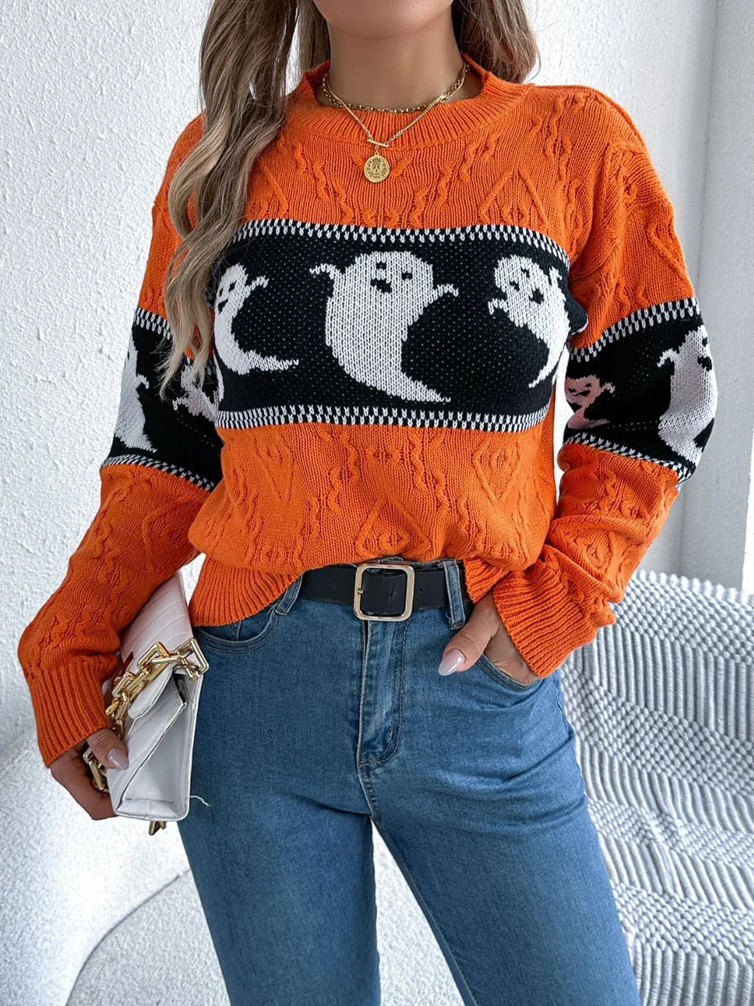Woman wearing an orange crew neck knit sweater with a ghost print pattern on the sleeves and a black and white striped band across the chest.