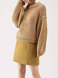 Woman posing with her arm raised, wearing a tan chunky knit sweater with balloon sleeves and a mock neckline, and a mustard yellow skirt.