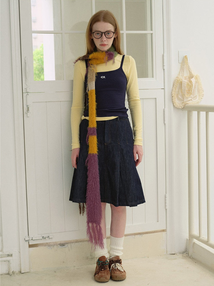 Full body shot of a woman wearing a yellow long-sleeve shirt, navy camisole, denim skirt, white socks, brown shoes, and a multi-colored striped fuzzy scarf.