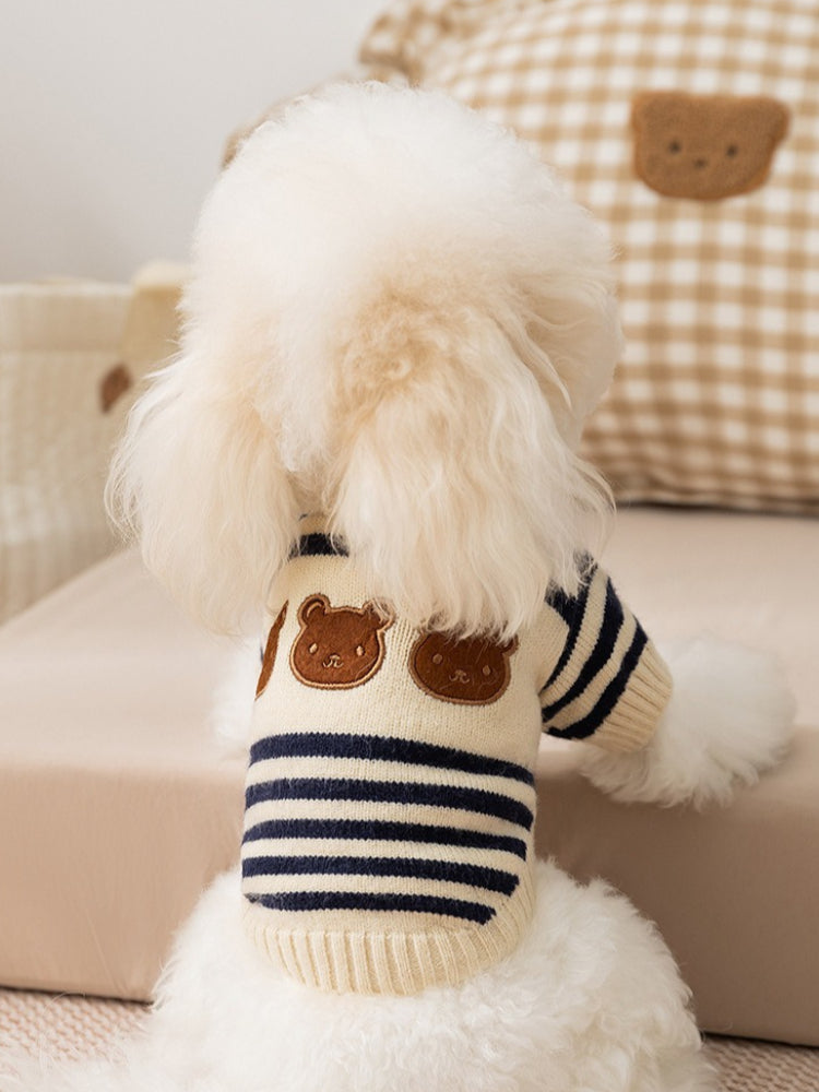 The back of a small white dog wearing a cream and navy blue striped knit sweater with two embroidered bear faces on the upper back.