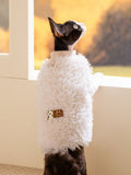 A black and white cat wearing a white faux fur coat with a decorative patch looking out a window.