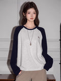 Woman wearing a light grey long-sleeve raglan shirt with navy blue sleeves and a small embroidered bird graphic, paired with khaki pants.