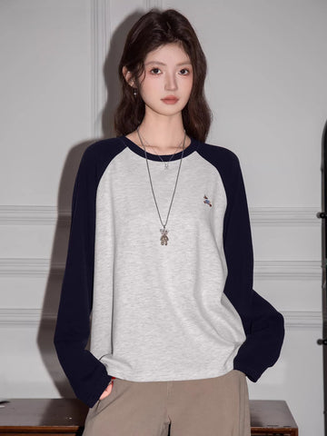 Woman wearing a light grey long-sleeve raglan shirt with navy blue sleeves and a small embroidered bird graphic, paired with khaki pants.
