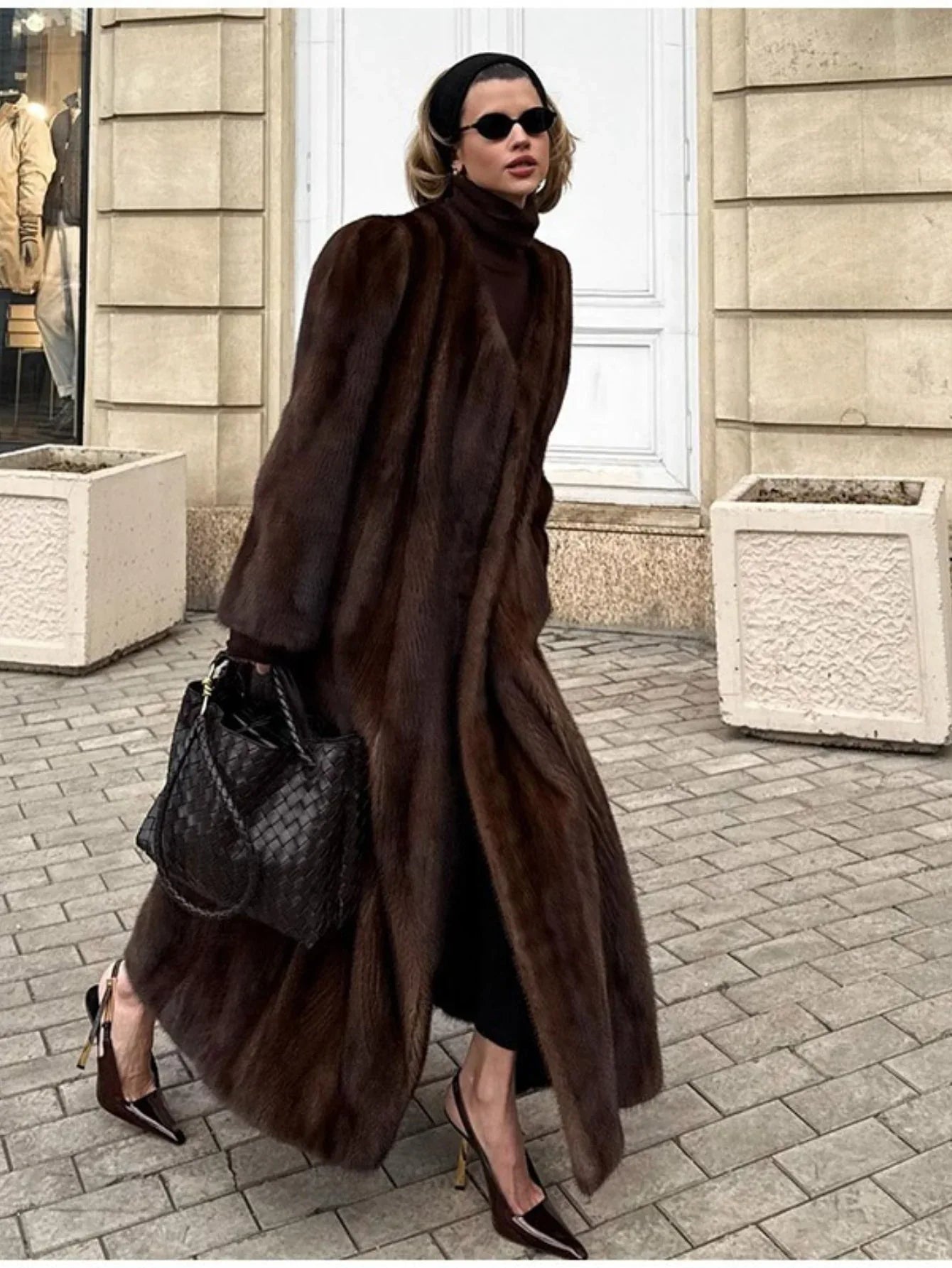 Woman wearing a long brown mink fur coat with a turndown collar, black turtleneck, black pants, black woven handbag, and brown pointed-toe heels walking outdoors.