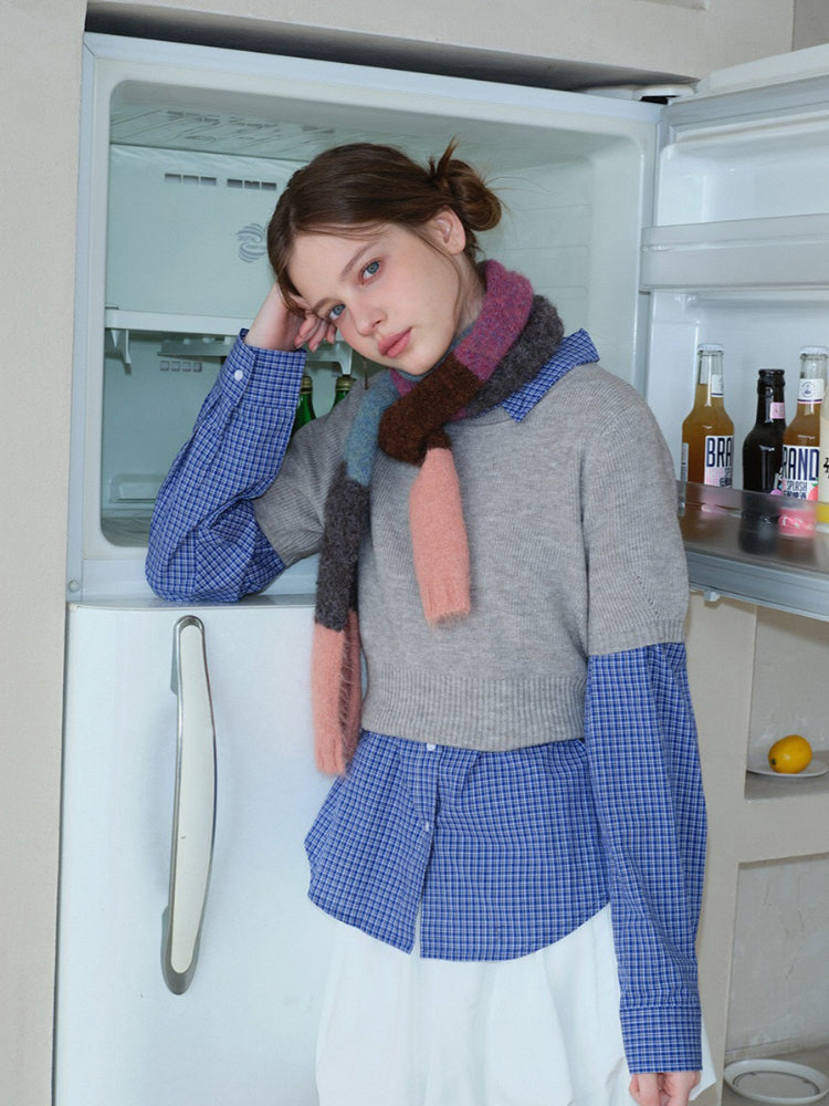 Young woman leaning against a refrigerator, styled in a casual layered outfit with a patterned scarf.
