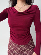Woman wearing a fitted burgundy long-sleeve top with a cowl neck and asymmetrical hem.