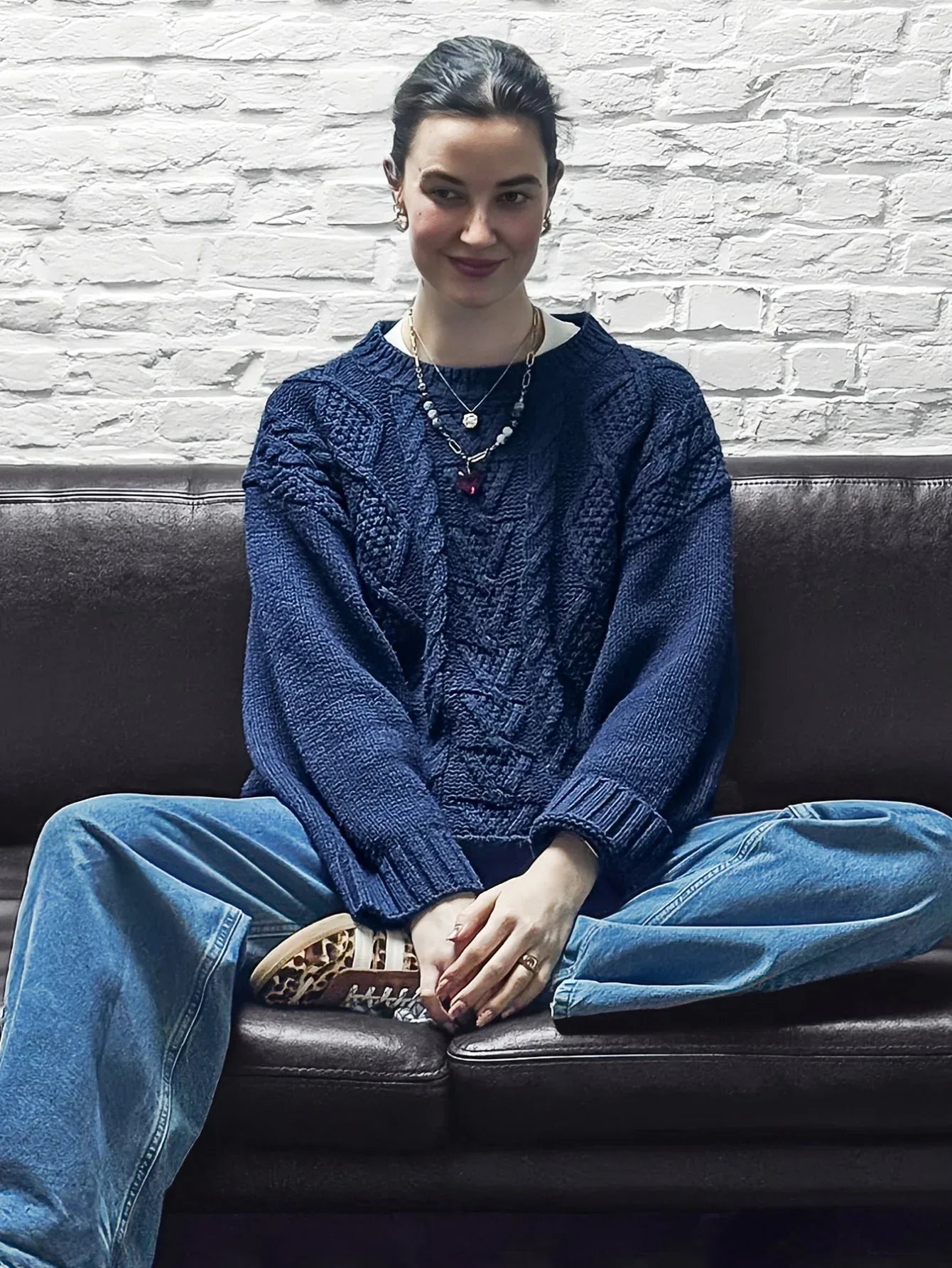 Woman sitting on a brown leather couch wearing a navy blue cable knit sweater and light wash jeans.