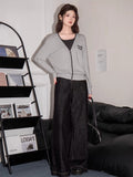 Woman wearing a grey V-neck knit cardigan with contrast trim and embroidered graphic detail with a dark top and wide-leg jeans
