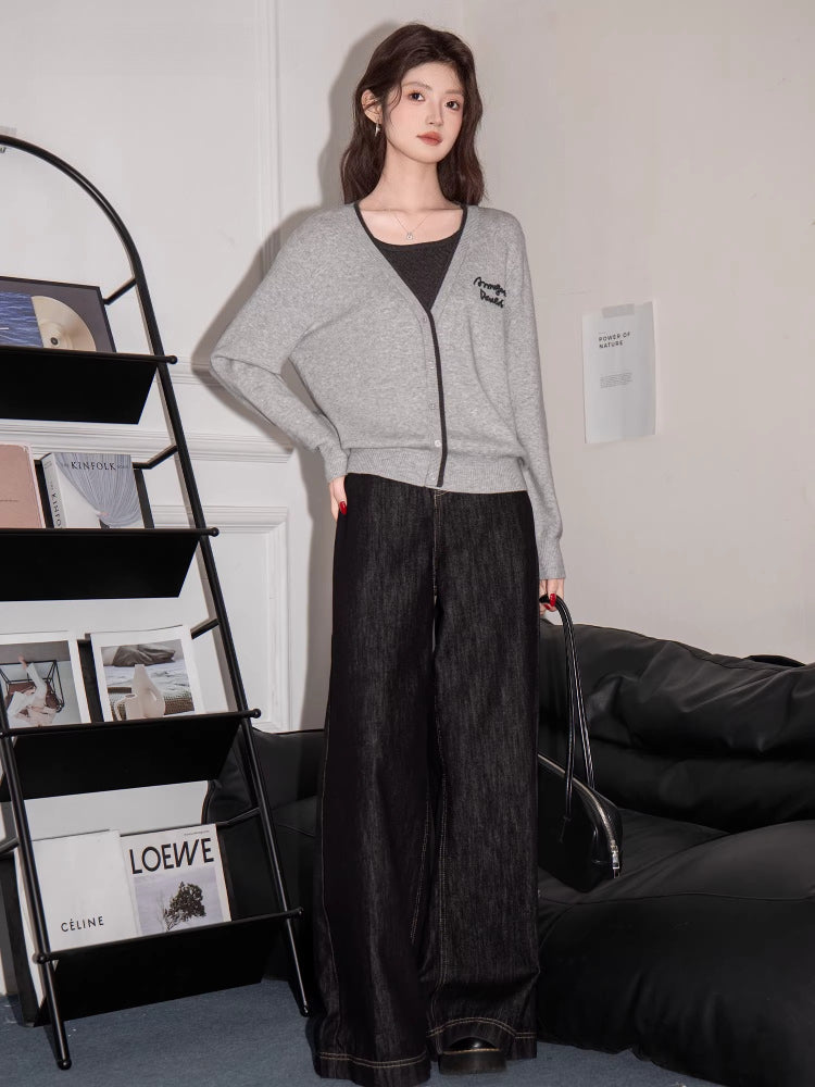 Woman wearing a grey V-neck knit cardigan with contrast trim and embroidered graphic detail with a dark top and wide-leg jeans