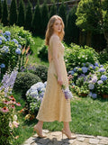 Woman walking in a garden wearing a sleeveless yellow jacquard floral midi dress with a round neckline and criss-cross back straps.